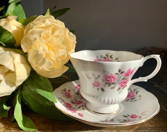 Puede incluir: Una taza de té y un platillo blancos con un patrón floral de rosas rosadas y hojas verdes. La taza de té tiene un borde festoneado y un asa. El platillo tiene un borde dorado.
