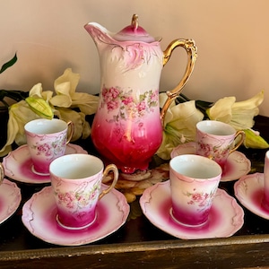 May include: A porcelain tea set featuring a tall pitcher and five matching cups with saucers. Each piece displays a white base with a vibrant magenta gradient at the bottom, decorated with delicate pink and green floral patterns and gold handles.