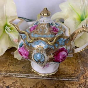 May include: A small, ornate porcelain teapot with a light blue background, pink roses, and gold accents. The teapot has a matching lid and a curved handle. The design is detailed and elegant, suitable for a tea party or display.