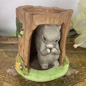 May include: A ceramic squirrel figurine inside a tree trunk shaped holder. The squirrel is grey and has a brown nose. The tree trunk is brown with green leaves and acorns.
