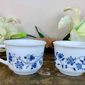 Puede incluir: Dos tazas de cerámica blanca con diseños florales azules. Las tazas tienen una forma simple y redondeada y un asa en el lateral.