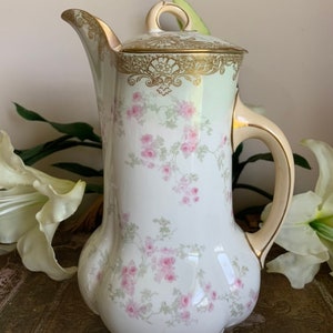 Puede incluir: Una cafetera de porcelana blanca con un patrón floral y detalles dorados. La cafetera tiene un cuerpo redondo y un pico curvo. La tapa tiene un borde dorado y un diseño floral.