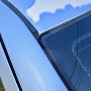 Puede incluir: Primer plano de la carrocería plateada y los embellecedores de goma negra de un coche. La imagen muestra la esquina del parabrisas y el borde del techo, destacando los detalles exteriores del vehículo.