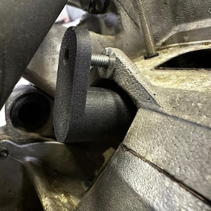 Puede incluir: Primer plano de un soporte metálico negro fijado a un componente metálico del motor con un perno. El soporte tiene una forma curva con un orificio de montaje. El componente del motor es de color gris apagado.