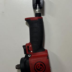 Puede incluir: Una llave de impacto roja y negra con un soporte de montaje negro adjunto. La herramienta tiene un mango negro con una parte superior roja y una cabeza negra. El soporte de montaje es negro y tiene un agujero para colgar.