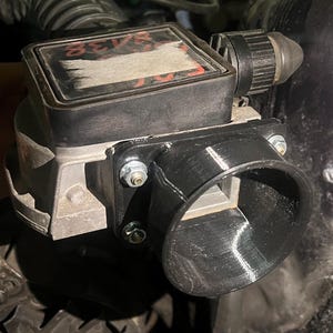 Puede incluir: Primer plano de una pieza de automóvil, probablemente un componente de admisión de aire. La pieza es principalmente negra y plateada, con una carcasa rectangular y una entrada cilíndrica. Sujeta con pernos, parece estar montada dentro de un compartimento del motor.