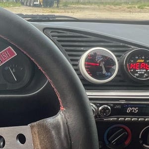 May include: Car dashboard interior with a black steering wheel, red stitching, and gauges. The dashboard has stickers with the words "DEATHWISH" and "TOUGHDREAMS". Gauges display readings, and a radio is visible.