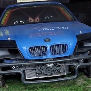 May include: A blue BMW race car with front-end damage. The car features a black roll cage, a large front bumper, and a radiator fan. Text on the bonnet reads "PERICOLO DI MORTE".