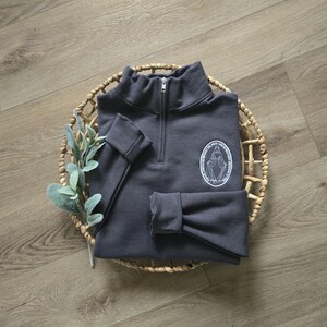 Puede incluir: Una sudadera gris carb&oacute;n con cremallera de un cuarto, cuello alto y mangas largas con pu&ntilde;os. En el pecho izquierdo, hay un gr&aacute;fico circular blanco que representa una figura con los brazos extendidos y el texto O MARY CONCEIVED WITHOUT SIN PRAY FOR US WHO HAVE RECOURSE TO THEE.