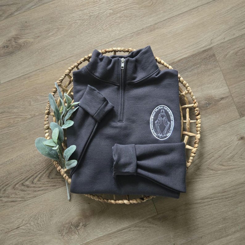 Puede incluir: Una sudadera gris carb&oacute;n con cremallera de un cuarto, cuello alto y mangas largas con pu&ntilde;os. En el pecho izquierdo, hay un gr&aacute;fico circular blanco que representa una figura con los brazos extendidos y el texto O MARY CONCEIVED WITHOUT SIN PRAY FOR US WHO HAVE RECOURSE TO THEE.