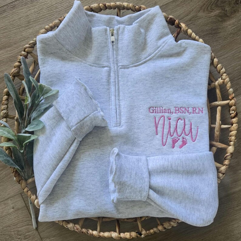K&ouml;nnte beinhalten: Ein hellgraues Sweatshirt mit Viertelrei&szlig;verschluss, bestickt mit dem Text "Gillian, BSN, RN" und "Nicu" in Rosa, sowie Babyf&uuml;&szlig;en. Das Sweatshirt hat einen gerippten Kragen und B&uuml;ndchen und wird in einem geflochtenen Korb pr&auml;sentiert.