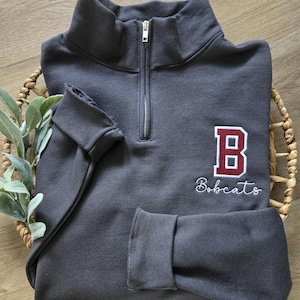 May include: A charcoal gray quarter-zip sweatshirt with a maroon and white embroidered "B Bobcats" design. The sweatshirt has a high collar, ribbed cuffs, and a silver zipper. The garment is folded in a woven basket.
