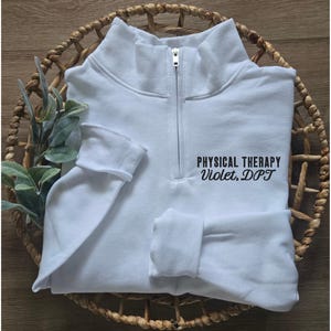 May include: A white quarter-zip sweatshirt with the text "PHYSICAL THERAPY Violet, DPT" in black. The sweatshirt is folded in a woven basket with green foliage.