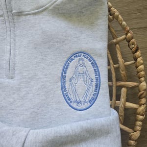 Puede incluir: Una sudadera gris jaspeado con cremallera de cuarto y un parche bordado azul de forma ovalada en el pecho. El bordado muestra una figura con el texto O MARY, CONCEIVED WITHOUT SIN, PRAY FOR US WHO HAVE RECOURSE TO THEE.