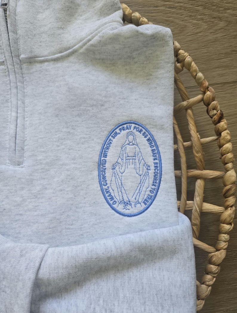 Puede incluir: Una sudadera gris jaspeado con cremallera de cuarto y un parche bordado azul de forma ovalada en el pecho. El bordado muestra una figura con el texto O MARY, CONCEIVED WITHOUT SIN, PRAY FOR US WHO HAVE RECOURSE TO THEE.