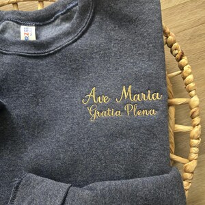 May include: A dark blue sweatshirt with the text "Ave Maria Gratia Plena" embroidered in gold.