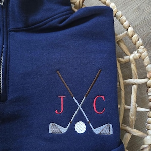 Peut inclure: Sweat-shirt bleu marine avec une fermeture &eacute;clair quart de zip. Il pr&eacute;sente un motif brod&eacute; de clubs de golf crois&eacute;s, d'une balle de golf et des lettres "J" et "C" en rouge. Le sweat-shirt est en mati&egrave;re douce.