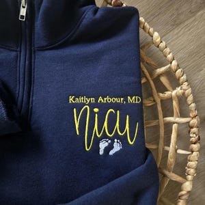 K&ouml;nnte beinhalten: Marineblauer Sweatshirt-Rei&szlig;verschluss mit dem Text "Kaitlyn Arbour, MD" und "NICU" in Gelb gestickt. Darunter befinden sich zwei kleine Babyf&uuml;&szlig;e in Hellblau. Das Sweatshirt liegt auf einem geflochtenen Korb.