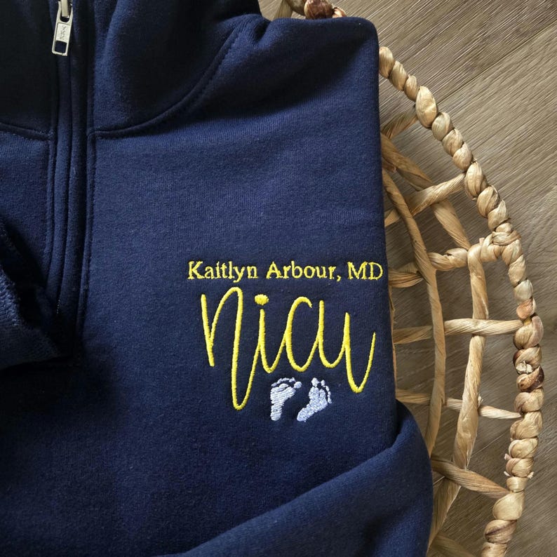 K&ouml;nnte beinhalten: Marineblauer Sweatshirt-Rei&szlig;verschluss mit dem Text "Kaitlyn Arbour, MD" und "NICU" in Gelb gestickt. Darunter befinden sich zwei kleine Babyf&uuml;&szlig;e in Hellblau. Das Sweatshirt liegt auf einem geflochtenen Korb.