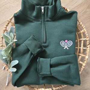 A forest green sweatshirt with white paddles, pink pickleball, and white initials.