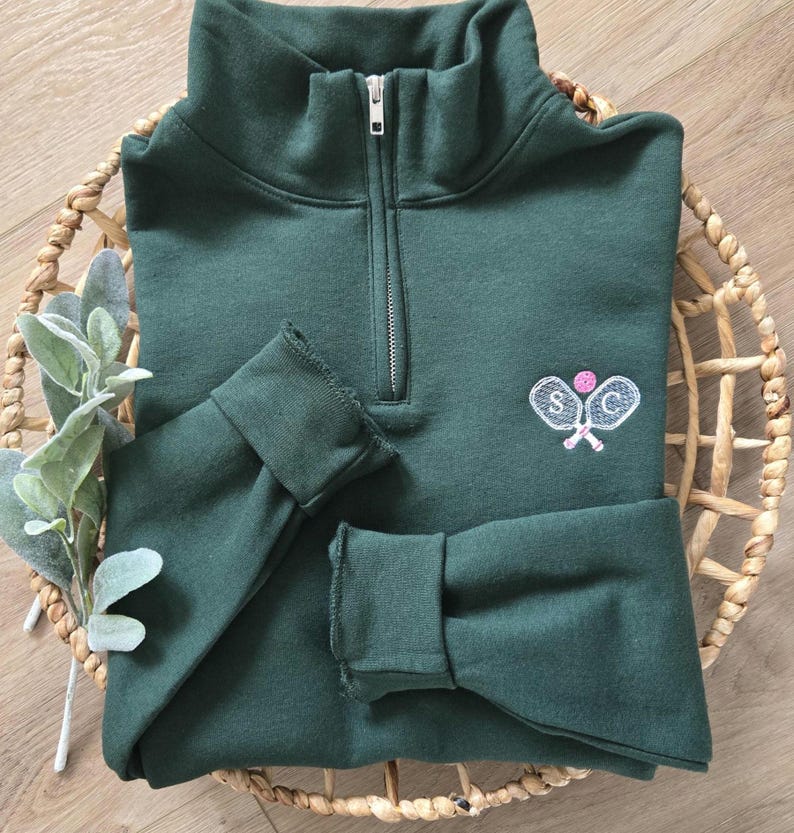 A forest green sweatshirt with white paddles, pink pickleball, and white initials.