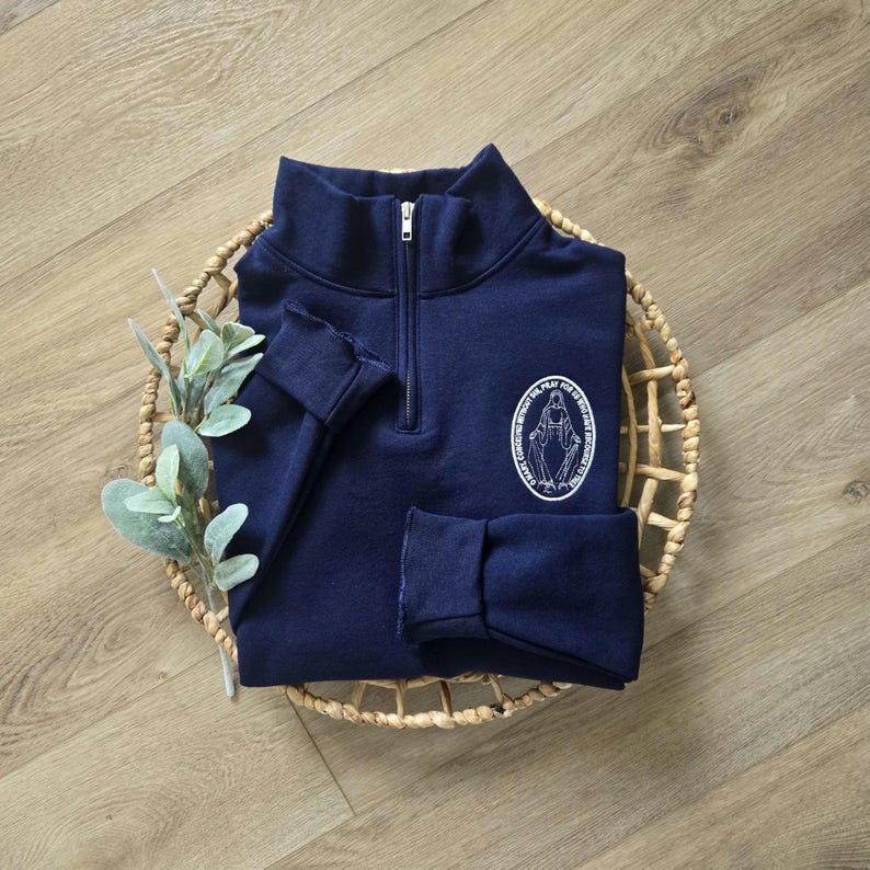Puede incluir: Una sudadera azul marino con cremallera de un cuarto y un gr&aacute;fico ovalado blanco en el pecho. El gr&aacute;fico muestra una figura con los brazos extendidos rodeada por el texto O MARY CONCEIVED WITHOUT SIN PRAY FOR US WHO HAVE RECOURSE TO THEE.