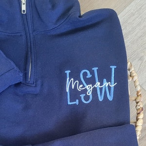 May include: A navy blue quarter-zip sweatshirt with a white embroidered name "Megan" and blue embroidered initials "L.S.W."