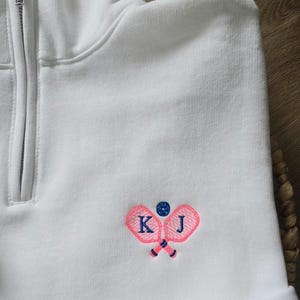 May include: White quarter-zip sweatshirt with pink tennis rackets and a blue tennis ball embroidered on the front. The rackets are crossed and the initials "K J" are embroidered on the rackets.