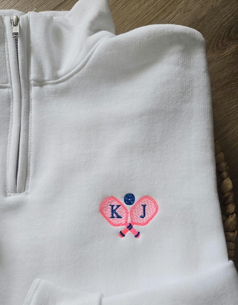 May include: White quarter-zip sweatshirt with pink tennis rackets and a blue tennis ball embroidered on the front. The rackets are crossed and the initials "K J" are embroidered on the rackets.