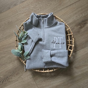May include: A gray quarter-zip sweatshirt with the name "Alexis" embroidered in cursive script. The sweatshirt is folded neatly in a woven basket with a sprig of greenery.