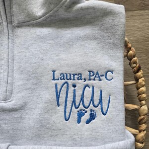 K&ouml;nnte beinhalten: Hellgraues Sweatshirt mit Viertelrei&szlig;verschluss und blau gesticktem Text "Laura, PA-C" und "NICU" mit Babyf&uuml;&szlig;en. Das Sweatshirt ist gefaltet und liegt auf einem geflochtenen Korb.