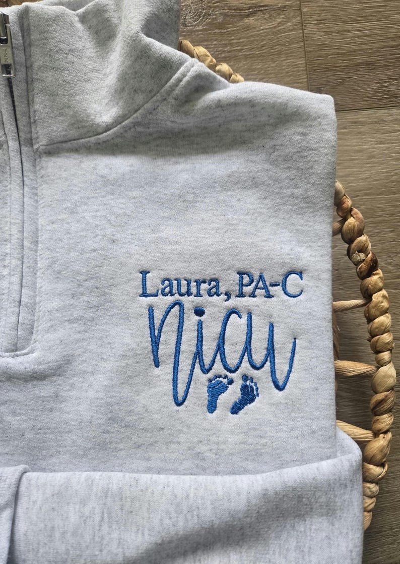 K&ouml;nnte beinhalten: Hellgraues Sweatshirt mit Viertelrei&szlig;verschluss und blau gesticktem Text "Laura, PA-C" und "NICU" mit Babyf&uuml;&szlig;en. Das Sweatshirt ist gefaltet und liegt auf einem geflochtenen Korb.