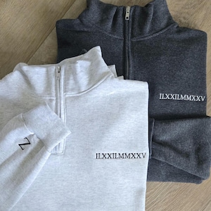 May include: Two quarter-zip sweatshirts, one gray and one dark gray, with the Roman numeral date "II.XXIII.MMXXV" embroidered on the right sleeve. The gray sweatshirt has the letter "Z" embroidered on the left sleeve.