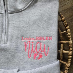 K&ouml;nnte beinhalten: Ein graues Sweatshirt mit Viertelrei&szlig;verschluss und dem gestickten Text "London, BSN, RN" &uuml;ber dem Wort "NICU" in rosa Schrift, zusammen mit zwei rosa Babyf&uuml;&szlig;en. Das Sweatshirt ist gefaltet und liegt in einem Weidenkorb.