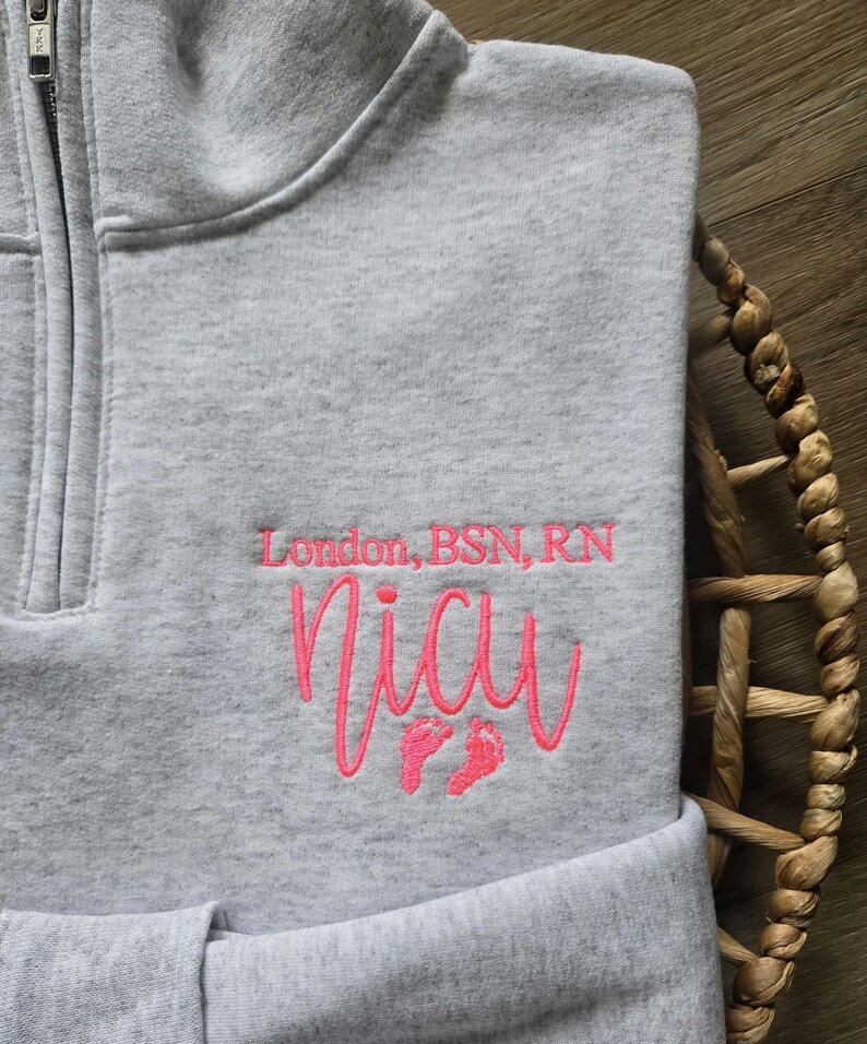 K&ouml;nnte beinhalten: Ein graues Sweatshirt mit Viertelrei&szlig;verschluss und dem gestickten Text "London, BSN, RN" &uuml;ber dem Wort "NICU" in rosa Schrift, zusammen mit zwei rosa Babyf&uuml;&szlig;en. Das Sweatshirt ist gefaltet und liegt in einem Weidenkorb.