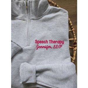Pu&ograve; includere: Felpa grigia con cerniera e il testo "Speech Therapy Jennifer, SLP" in rosa.