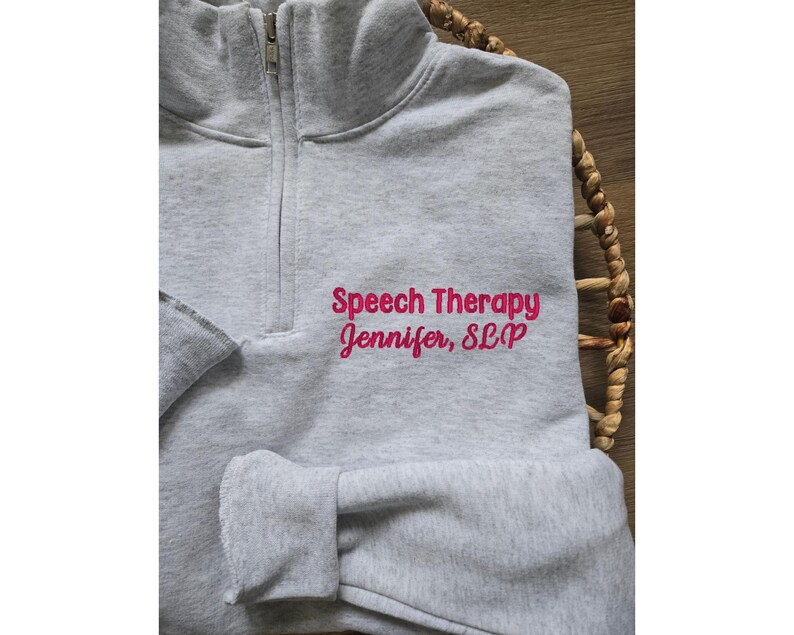 Pu&ograve; includere: Felpa grigia con cerniera e il testo "Speech Therapy Jennifer, SLP" in rosa.