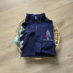 Puede incluir: Una sudadera azul marino con cremallera de un cuarto de largo que presenta un bordado de Jesús con rayos de luz rojos y azules que emanan del pecho, junto al texto bordado "Jesus, I trust in You" en una fuente cursiva.