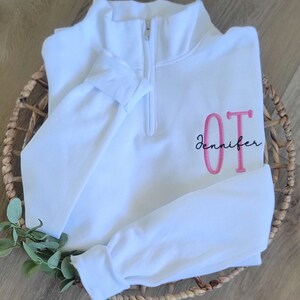 May include: White quarter-zip sweatshirt with a pink embroidered "OT" and the name "Jennifer" in black script.