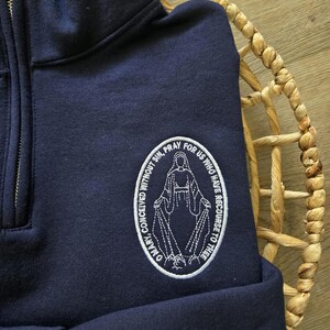 Puede incluir: Una sudadera azul marino con cremallera corta y un parche ovalado bordado en blanco en el pecho. El parche muestra una figura con los brazos extendidos rodeada por el texto, O MARY, CONCEIVED WITHOUT SIN, PRAY FOR US WHO HAVE RECOURSE TO THEE.