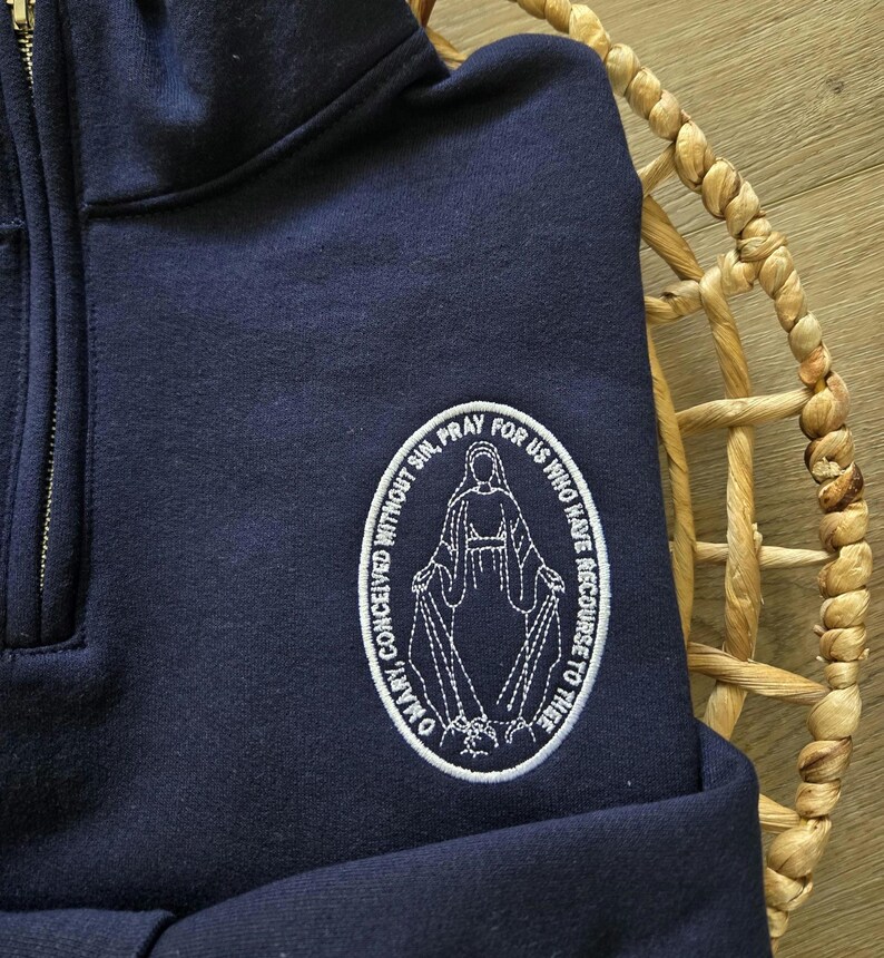 Puede incluir: Una sudadera azul marino con cremallera corta y un parche ovalado bordado en blanco en el pecho. El parche muestra una figura con los brazos extendidos rodeada por el texto, O MARY, CONCEIVED WITHOUT SIN, PRAY FOR US WHO HAVE RECOURSE TO THEE.