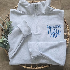 K&ouml;nnte beinhalten: Hellgraues Sweatshirt mit Viertelrei&szlig;verschluss und der gestickten Aufschrift "Laura, PA-C" &uuml;ber "NICU" in blauer Schrift, zusammen mit winzigen blauen Fu&szlig;abdr&uuml;cken. Das Sweatshirt ist in einem geflochtenen Korb gefaltet.