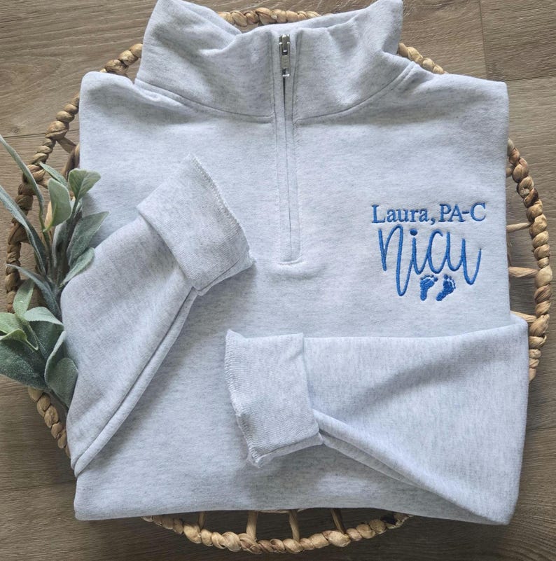 K&ouml;nnte beinhalten: Hellgraues Sweatshirt mit Viertelrei&szlig;verschluss und der gestickten Aufschrift "Laura, PA-C" &uuml;ber "NICU" in blauer Schrift, zusammen mit winzigen blauen Fu&szlig;abdr&uuml;cken. Das Sweatshirt ist in einem geflochtenen Korb gefaltet.