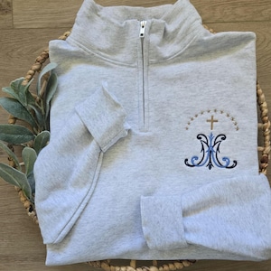 May include: A gray quarter-zip sweatshirt with a blue and gold embroidered design featuring a cross and the letters "M" and "M".