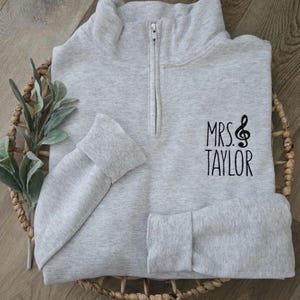 May include: A gray quarter-zip sweatshirt with a black embroidered design that says "MRS. TAYLOR" with a treble clef.