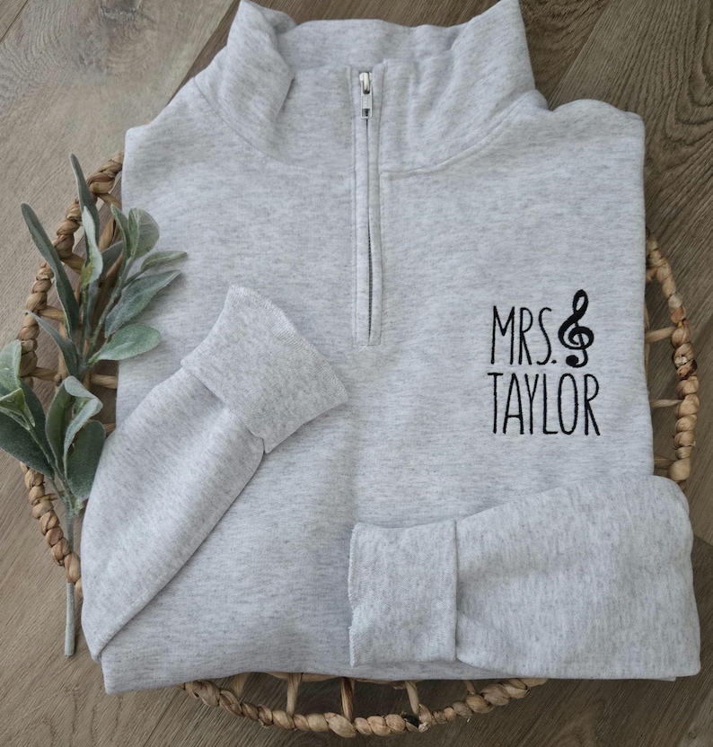 May include: A gray quarter-zip sweatshirt with a black embroidered design that says "MRS. TAYLOR" with a treble clef.
