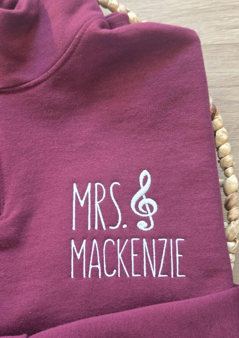 May include: Burgundy colored sweatshirt with white embroidery that reads "MRS. &" with a treble clef symbol and "MACKENZIE".