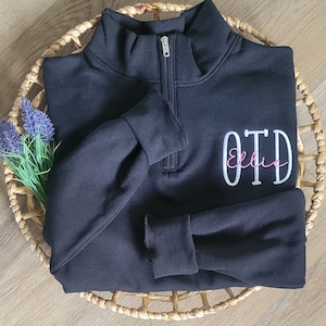 May include: Black quarter-zip sweatshirt with a white embroidered monogram "OTD" and the name "Ellie" in pink script.