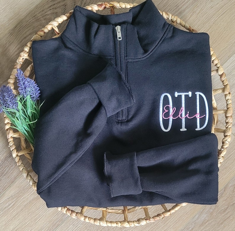 May include: Black quarter-zip sweatshirt with a white embroidered monogram "OTD" and the name "Ellie" in pink script.