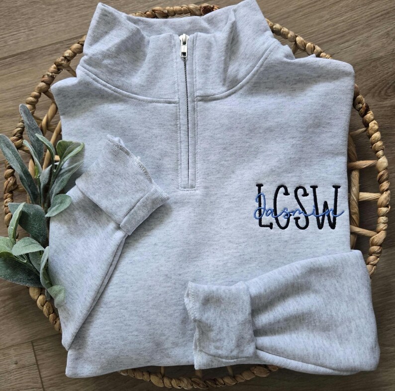 May include: A grey quarter-zip sweatshirt with a black embroidered design that says "LCSW" in a cursive font.